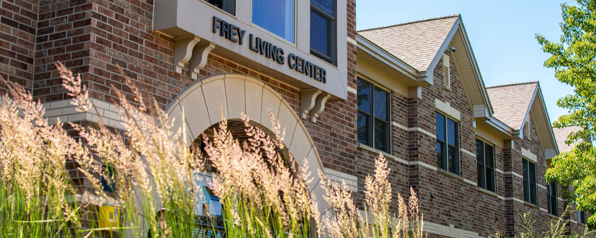 Brick building named Frey Living Center, with tall ornamental grasses in the foreground and a clear, sunny sky above, creating a welcoming feel.